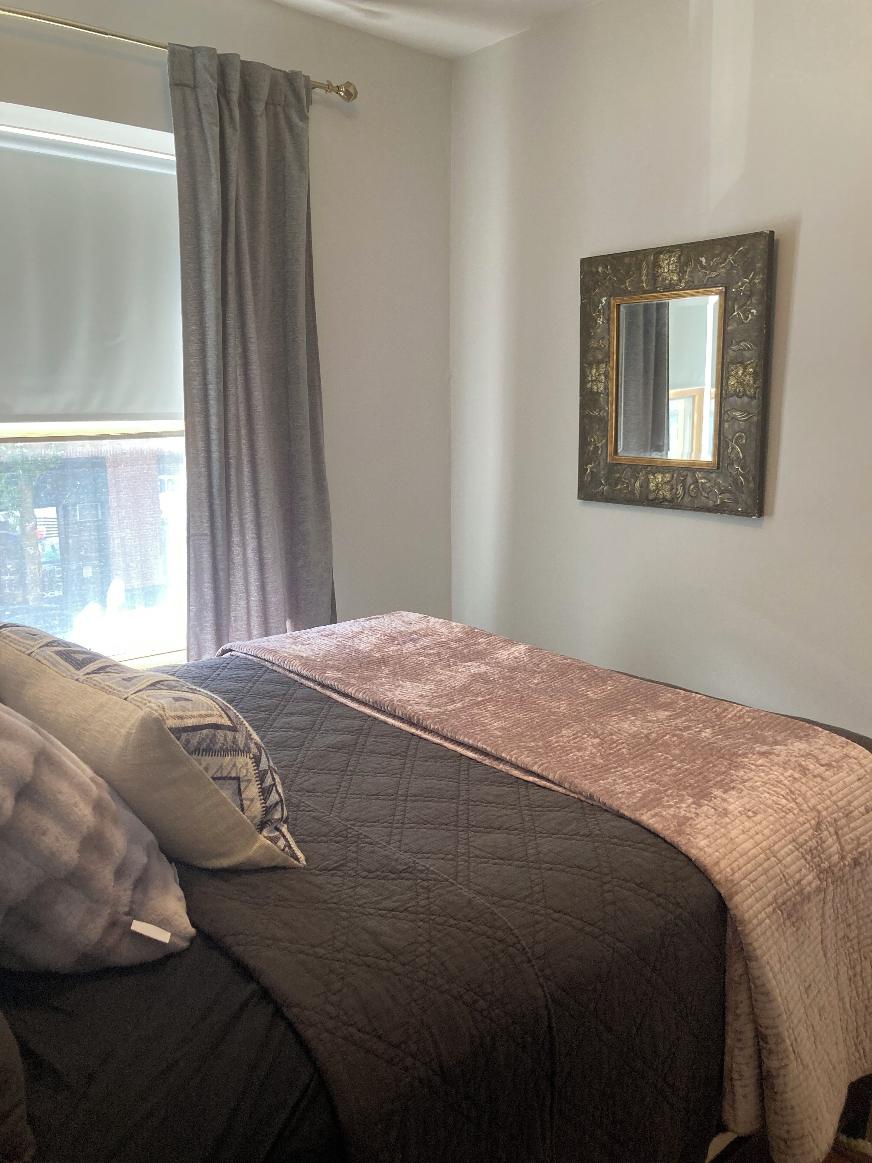 A bedroom with a bed, a window with curtains, and a wall-mounted mirror.