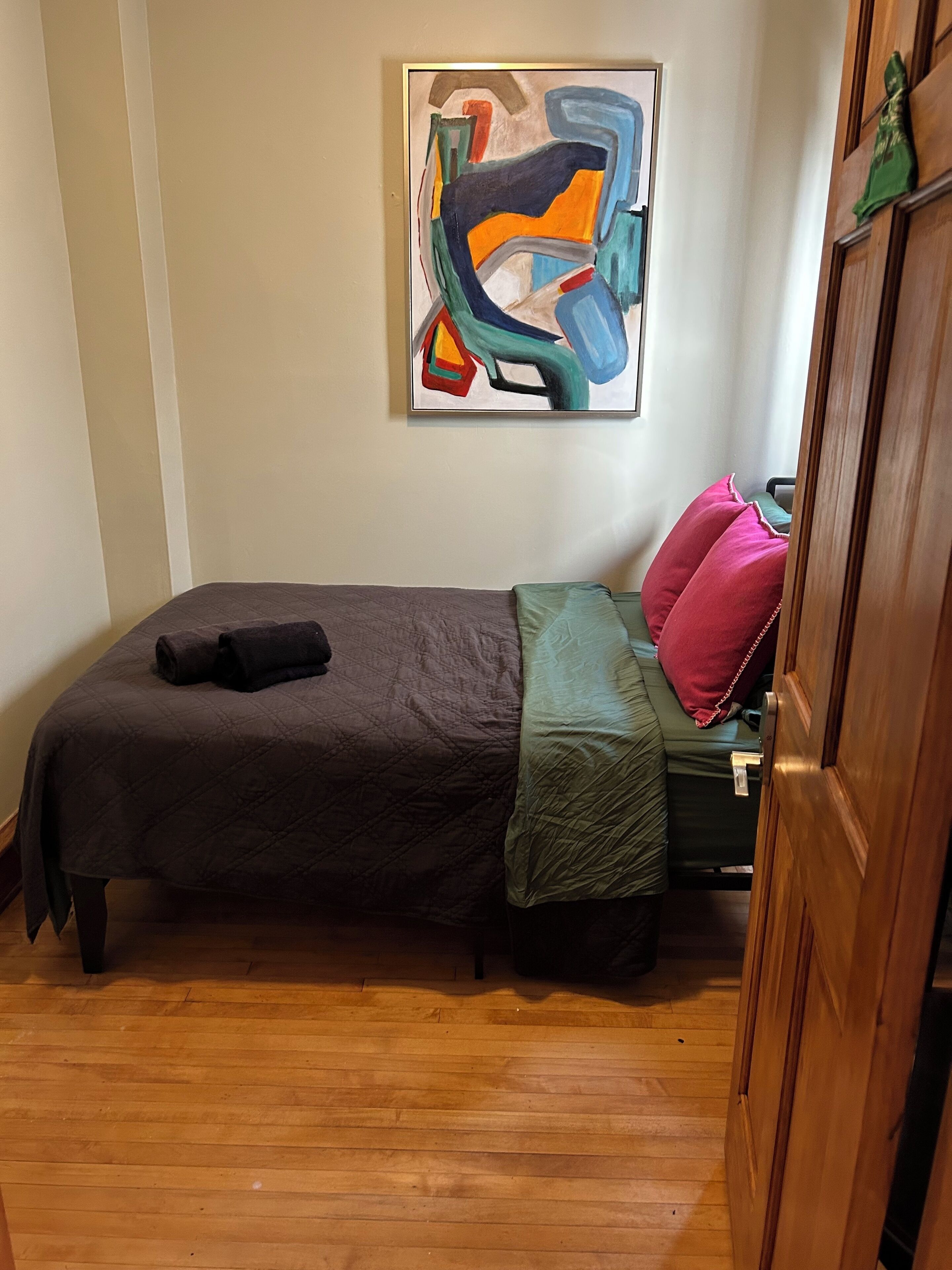 A small, neatly made bed with a dark-colored bedspread and pillows, a green throw blanket, and a colorful abstract painting on the wall.