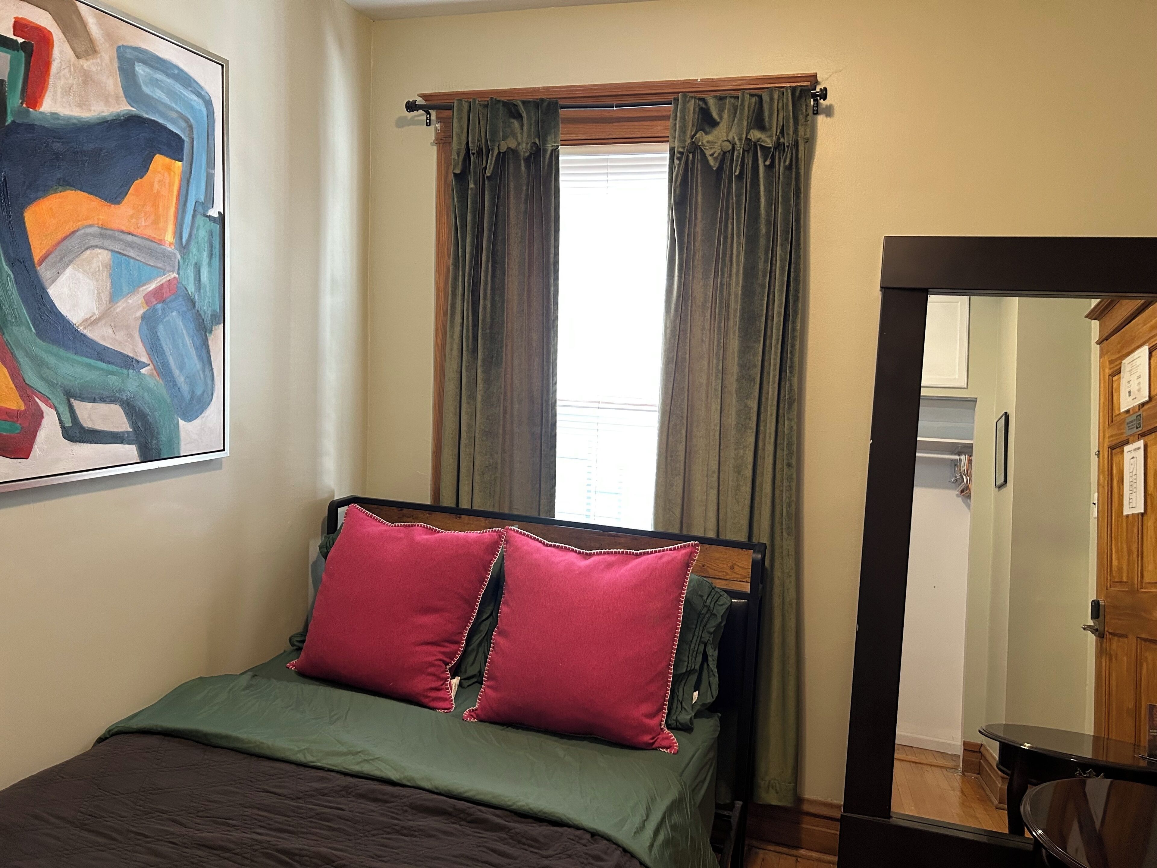 A bedroom with a bed, two pillows, a painting, a window with curtains, and a mirror.