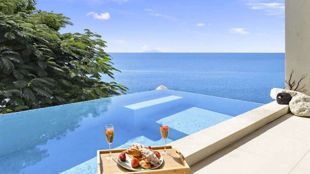 Ein Pool ohne Ende mit freiem Blick auf das Meer, ein Holztisch mit Speisen und Getränken sowie eine Lounge mit Meerblick.