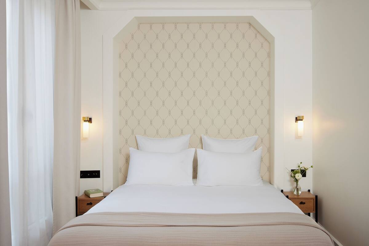 A neatly made bed with white linens and two pillows, a bedside table with a book and a vase of flowers, and a wall with a geometric pattern.