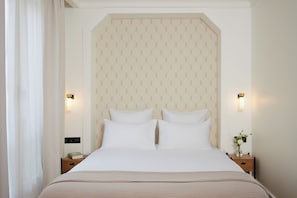 Una cama bien hecha con sábanas blancas, cabecera con un diseño geométrico, lámparas de noche y una mesita de noche con un libro y un florero.
