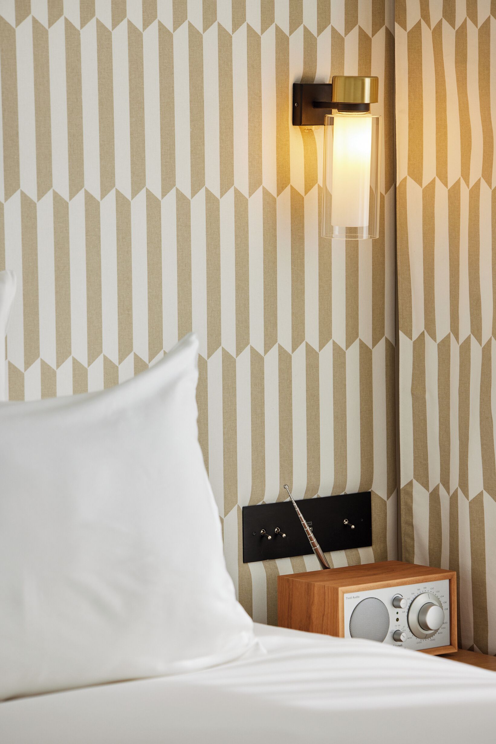 Ett hotellrum med randig tapet, en sänglampa, ett träbord vid sängen och en radio.