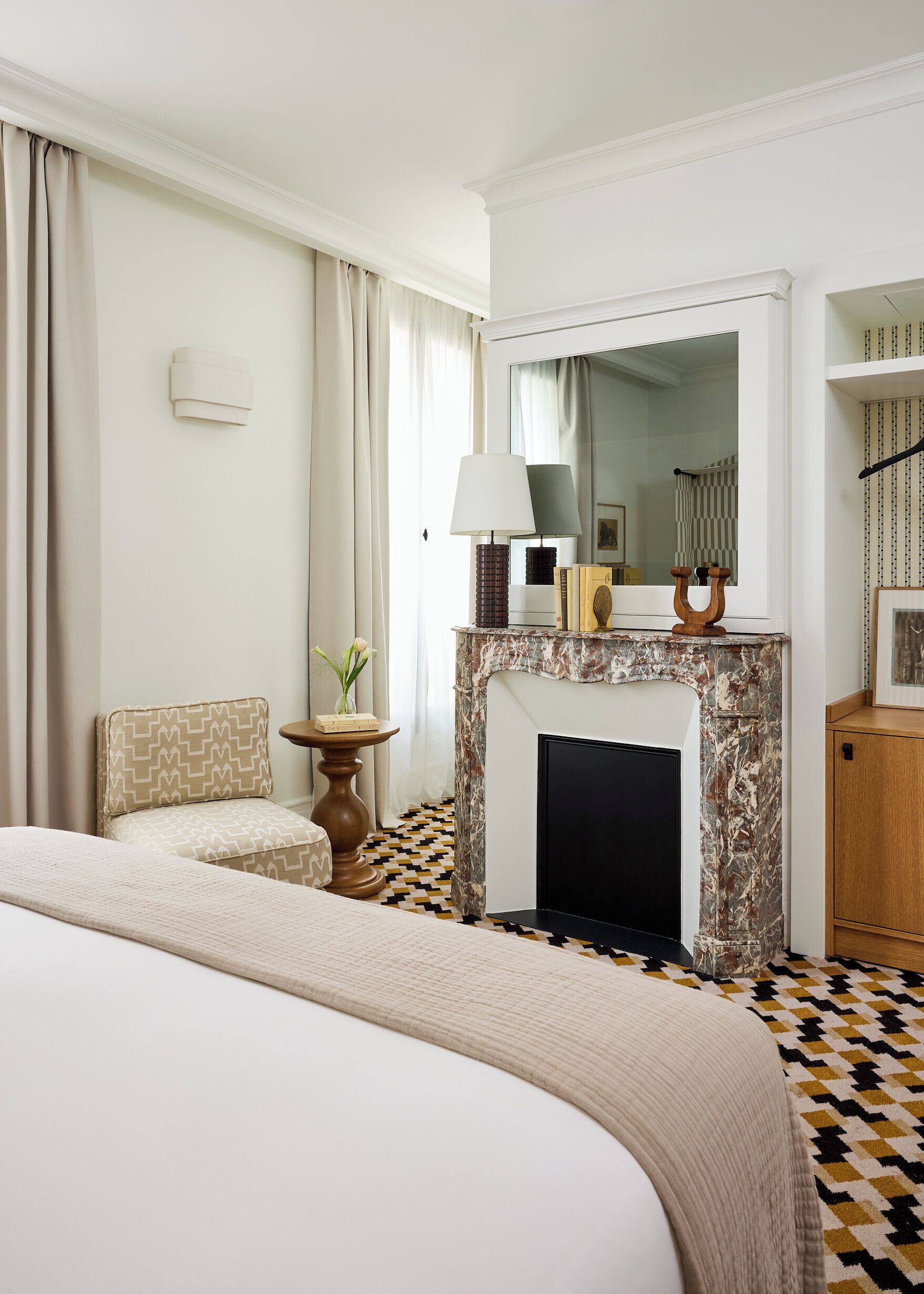 A hotel room with a bed, a fireplace, a mirror, a side table with a lamp, and a patterned floor.