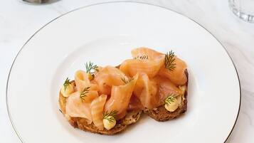 En tallrik med rökt lax på rostat bröd, en fruktskål med jordgubbar och bananer och ett glas vatten på ett marmorbord.