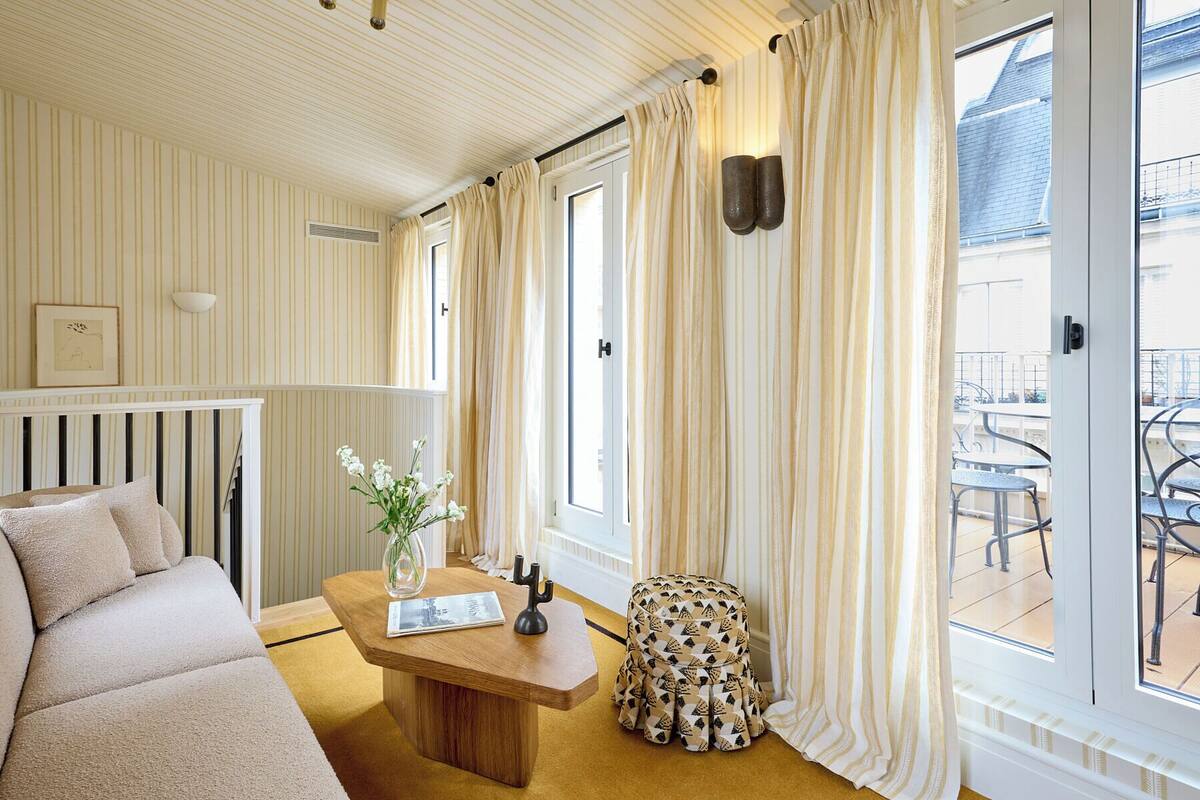A room with striped wallpaper, a wooden bench, a small table with a vase and a book, and a patterned stool.