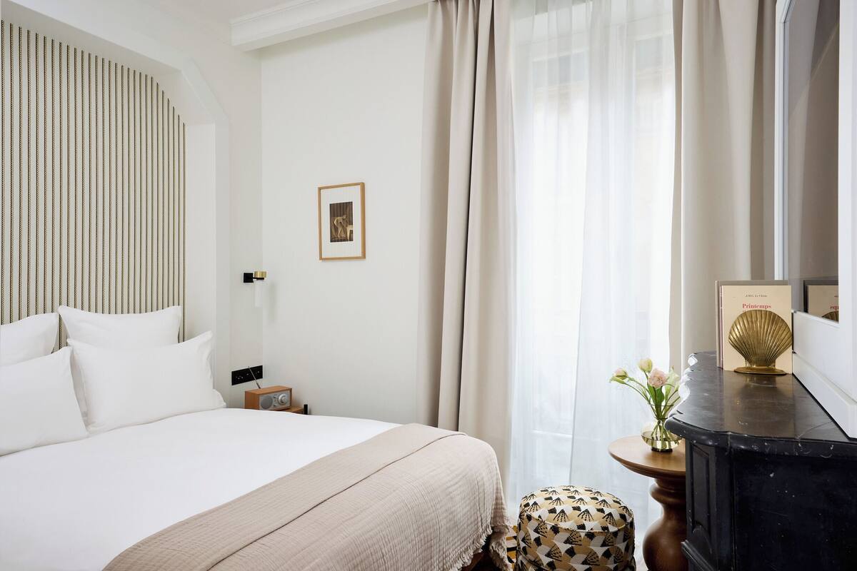 A hotel room with a bed, a bedside table with a lamp, a small table with a vase of flowers, and a mirror on the wall.