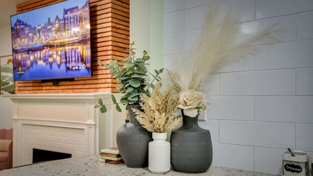 A kitchen counter with three vases containing plants and a decorative item, a television mounted on a wooden wall, and a tiled backsplash. - Stylish 2BRM APT: Putting Green: 1mi Masters: 2mi Medical Center (Augusta)