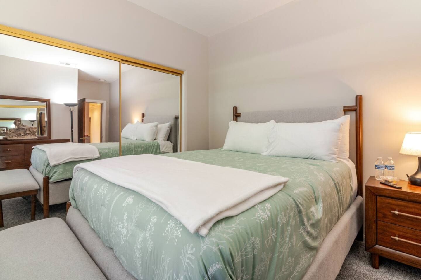 A hotel room with a large bed, a bedside table with a lamp, a mirror, and a small desk with a chair.