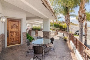 A covered patio with a table and chairs, a wooden door with a crest, and a view of a road and palm trees. - Mountain View Oasis  AC, Pool, Pet & Family Friendly (Hemet)