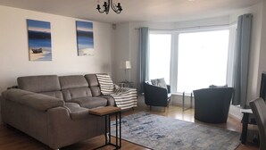 Ein modernes Wohnzimmer mit einem grauen Sofa, einem kleinen Beistelltisch, einem blauen Teppich und einem großen Fenster mit Vorhängen.