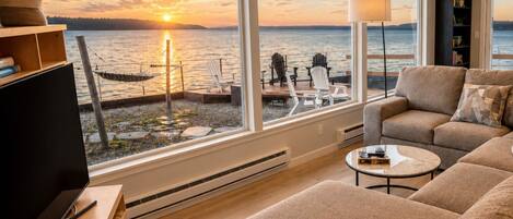 A modern living room with a large window overlooking a body of water at sunset, a sofa, a coffee table, and a television.