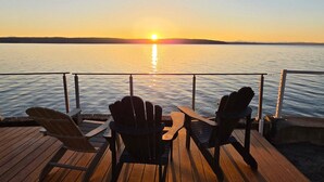 A sunset view from a wooden deck with two Adirondack chairs facing a calm body of water. - Hot Tub by the Water | Sunset Deck | Beach Access (Greenbank)