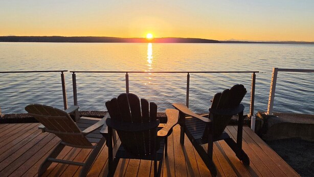 Zonsondergang zicht vanaf een houten terras met twee Adirondack-stoelen gericht op een kalm wateroppervlak.