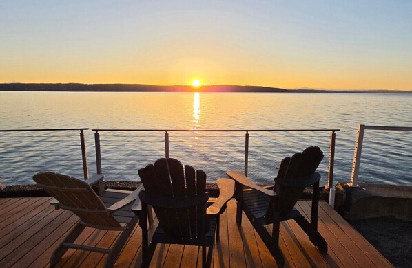 Une vue sur le coucher de soleil depuis une terrasse en bois, avec deux fauteuils Adirondack donnant sur une étendue d’eau calme.