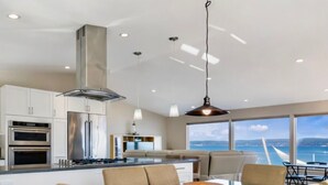 Une cuisine moderne dotée d’appareils électroménagers en acier inoxydable, une table à manger en verre avec quatre chaises, et une vue sur la mer grâce à de grandes fenêtres.