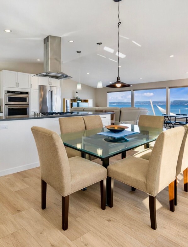 Een moderne keuken met roestvrijstalen apparatuur, een glazen eettafel met vier stoelen, en uitzicht op de zee door grote ramen.