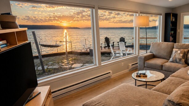 A living room with a large window overlooking a body of water at sunset, a television, and a sofa. - Hot Tub by the Water | Sunset Deck | Beach Access (Greenbank)