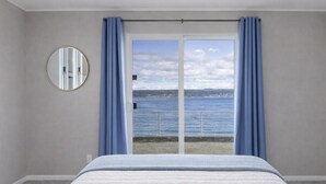 A bedroom with a bed, a round mirror, and a view of the sea through a sliding glass door. - Hot Tub by the Water | Sunset Deck | Beach Access (Greenbank)