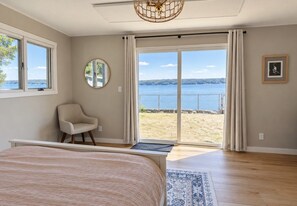 A bedroom with a large bed, a chair, a round mirror, and a view of the beach through a sliding glass door.