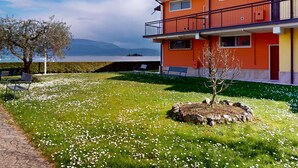 Un edificio de dos pisos con un balcón, un césped con flores blancas y un árbol.