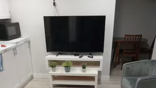 Un salon moderne avec un téléviseur à écran plat posé sur un meuble blanc, deux fauteuils gris, un tapis à motifs et un petit coin cuisine équipé d’un four à micro-ondes.