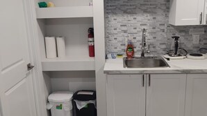 Une cuisine moderne avec des armoires blanches, un dosseret carrelé et un évier en acier inoxydable. Il y a une horloge au mur et un extincteur sur l’étagère.