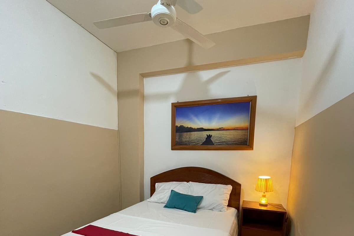 A neatly made bed with a red runner, a nightstand with a lamp, a ceiling fan, and a framed picture of a sunset over water on the wall.