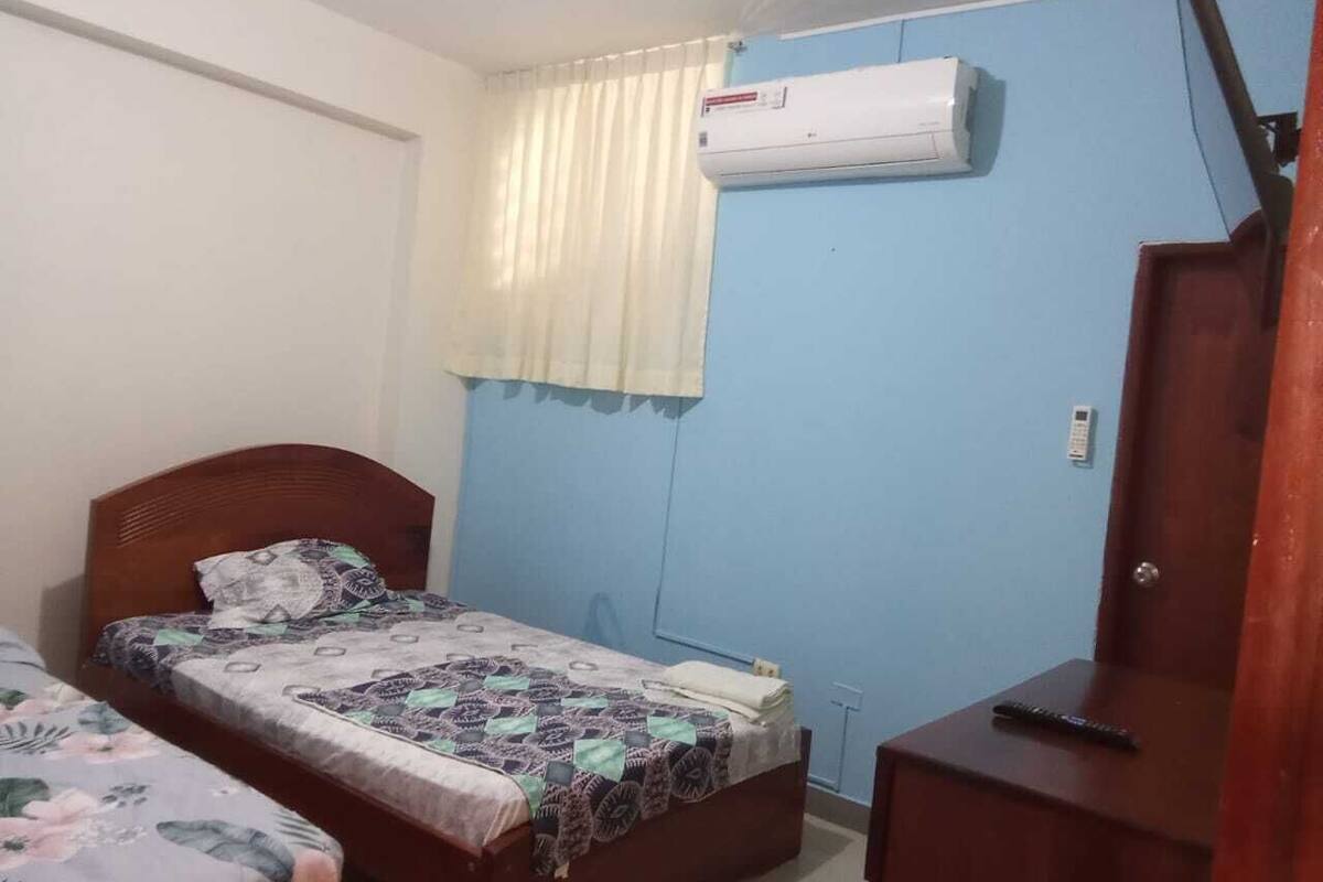 A bedroom with two single beds, a wooden wardrobe, a bedside table, an air conditioning unit, and a window with curtains.