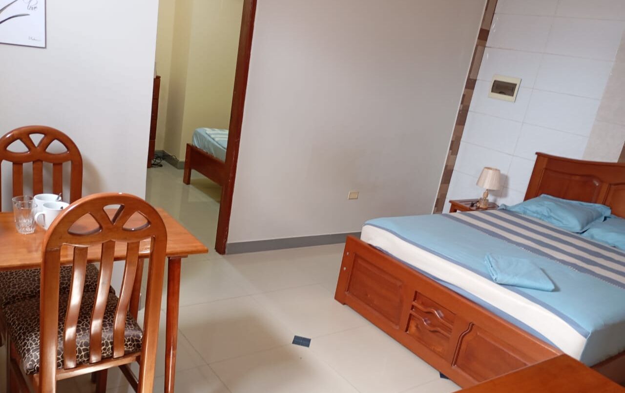 A bedroom with a wooden bed, a bedside table with a lamp, and a chair at a small table with a cup and glass.