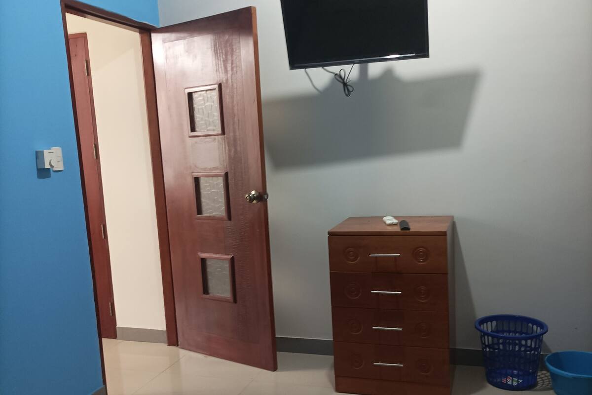 A bedroom with a bed, a wooden dresser, a television on the wall, and a blue door.