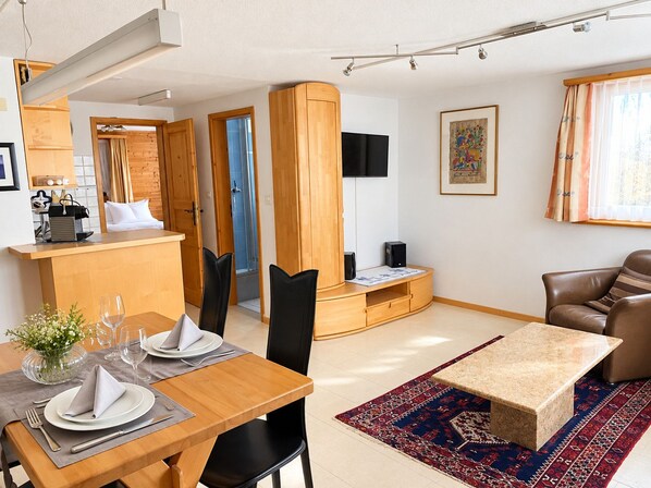 A modern living room with a dining area, a flat-screen TV mounted on the wall, and a large window with curtains.