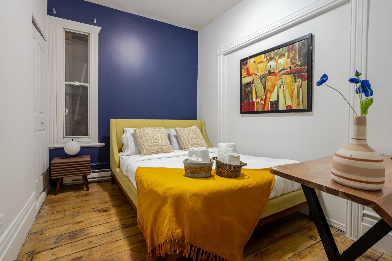 Een slaapkamer met een bed, een nachtkastje, een raam en een schilderij aan de muur.