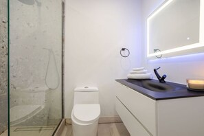 Une salle de bain moderne dotée d’une douche à parois de verre, de toilettes blanches, d’un lavabo sur un plan de toilette foncé et d’un miroir avec éclairage intégré.