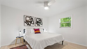 Ein Schlafzimmer mit einem Bett, einem Deckenventilator, einem Fenster mit Jalousien und abstrakter Kunst an der Wand.