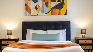 A neatly made bed with a dark headboard, two bedside tables with lamps, and a colorful abstract painting on the wall. - The Metro Stay – Vibrant 3BR in Gay Village (Montréal)