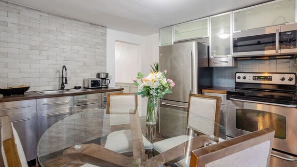 A modern kitchen with stainless steel appliances, a glass dining table, and a vase of flowers. - The Metro Stay – Vibrant 3BR in Gay Village (Montréal)