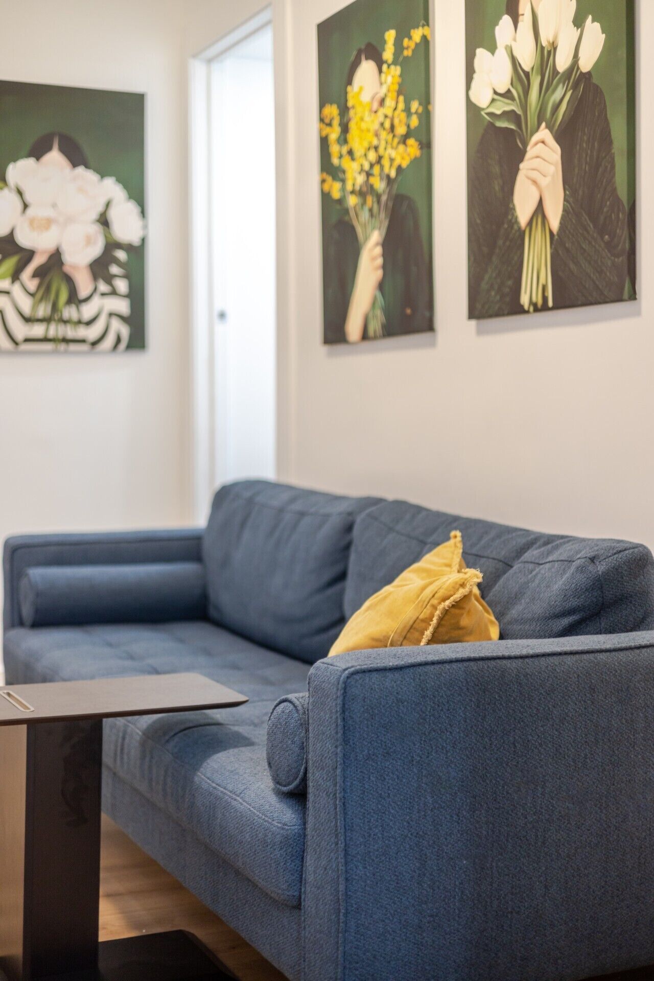 Un canapé bleu avec un coussin jaune, une petite table et trois œuvres d’art encadrées accrochées au mur.