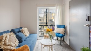 A modern living room with a blue sofa, a small round table with a vase of flowers, a blue armchair, and a sliding glass door leading to an outdoor balcony. - The Prestige 3BR| Griffintown Views (Montréal)