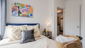 A bedroom with a bed, pillows, a bedside lamp, a wall art piece, and a wicker chair. - The Prestige 3BR| Griffintown Views (Montréal)