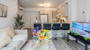 Een moderne woonkamer met een bank, een bijzettafeltje met bloemen, een televisie en een keuken op de achtergrond.