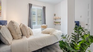 Een slaapkamer met een bed, een raam met gordijnen, een nachtkastje met een plant en een deur.