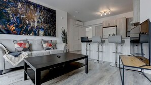 Ein modernes Wohnzimmer mit einer Couch, einem Couchtisch und Barhocker. An der Wand hängt ein großes abstraktes Gemälde, und durch eine offene Tür ist eine Küchenzeile zu sehen.