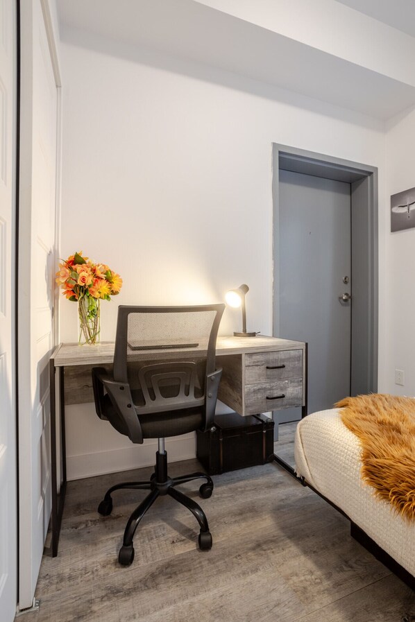 Une chambre à coucher avec un lit, un bureau avec une chaise, une commode, une porte et un vase avec des fleurs.