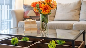 A glass-top coffee table with a stack of books, a vase with orange flowers, and small green plants. - Chic and Spacious 3 Bedrooms in Griffintown (Montréal)