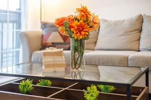 A glass-top coffee table with a stack of books, a vase with orange flowers, and small green plants.