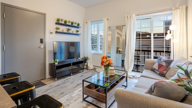 A modern living room with a flat-screen TV, a glass coffee table, a sofa, and a dining area with stools. - Chic and Spacious 3 Bedrooms in Griffintown (Montréal)