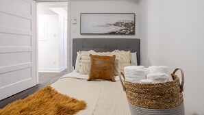 A bedroom with a grey headboard, a white door, a framed landscape painting, a brown throw pillow, a white rug, and a woven basket with towels. - Amazing 4 Bedroom Unit in Downtown (Montréal)