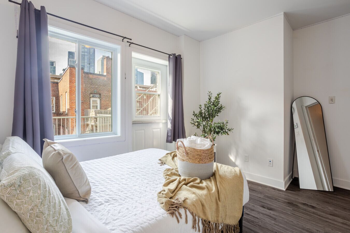 A bedroom with a bed, a window with curtains, a plant, a mirror, and a view of buildings outside.