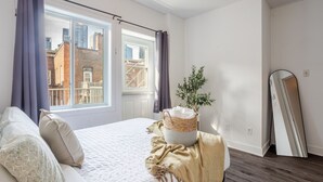 A bedroom with a bed, a window with curtains, a plant, a mirror, and a view of buildings outside. - Luxurious 4 BR Suite in Griffintown (Montréal)